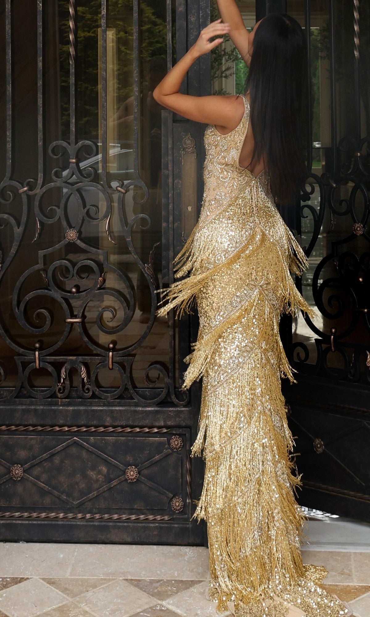 A woman wearing the Jovani 48521 long prom dress with a shimmering sequined beaded bodice stands with her back to the camera, raising her arms in front of ornate black metal doors.